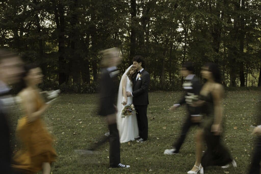 Bride and groom share an embrace in a wooded clearing at The Pick Inn, as their wedding party blurs past them in motion—capturing stillness amid joyful chaos