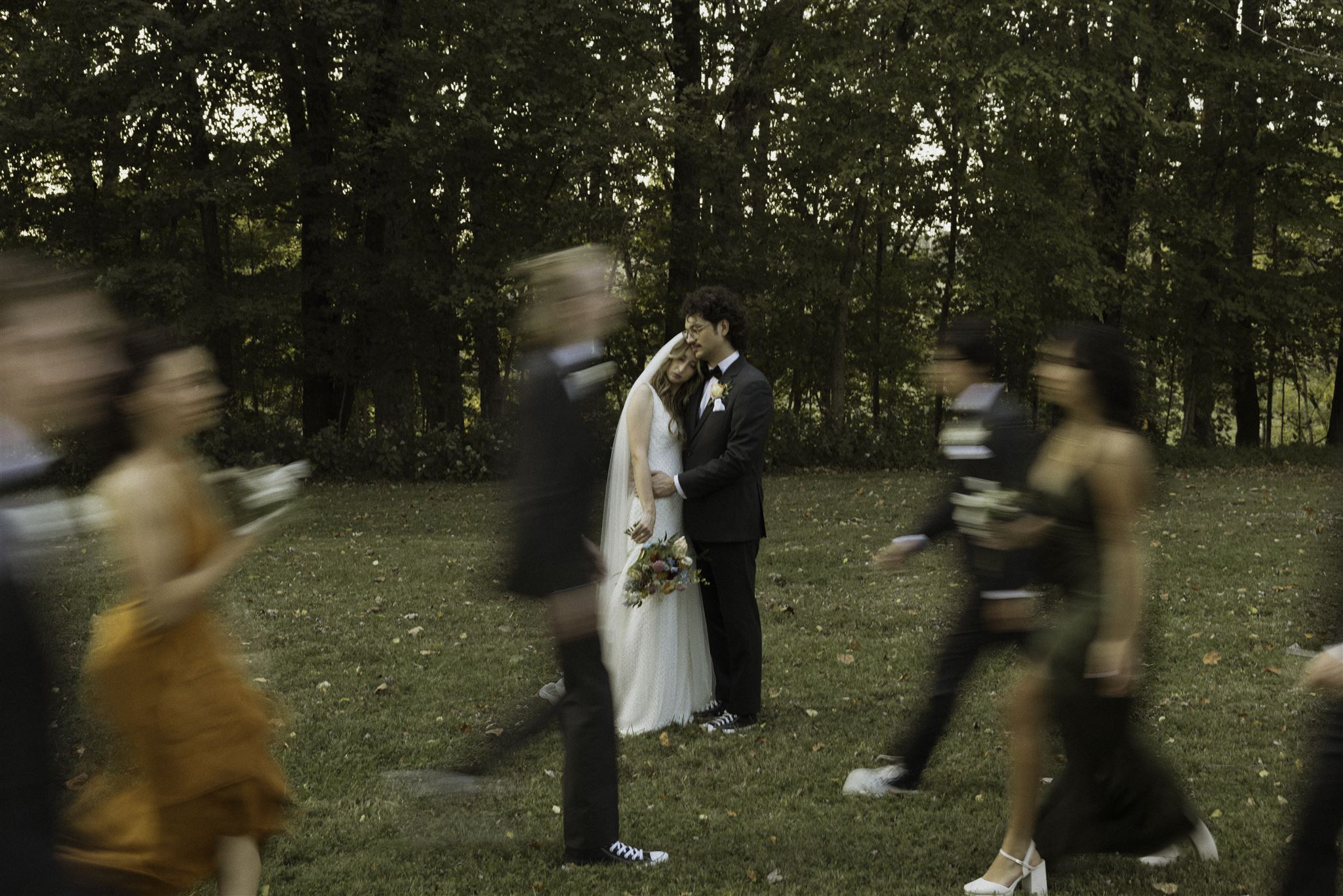 Bride and groom share an embrace in a wooded clearing at The Pick Inn, as their wedding party blurs past them in motion—capturing stillness amid joyful chaos